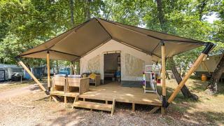 Tienda safari con terraza de madera y asientos exteriores, ubicada en un bosque rodeado de árboles verdes.