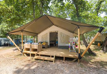 Tenda safari con veranda in legno e seduta esterna, immersa in un campeggio tra alberi verdi e natura.
