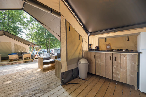 Binnenzicht van een moderne safaritent met kitchenette, houten meubelen en nog een tent buiten.