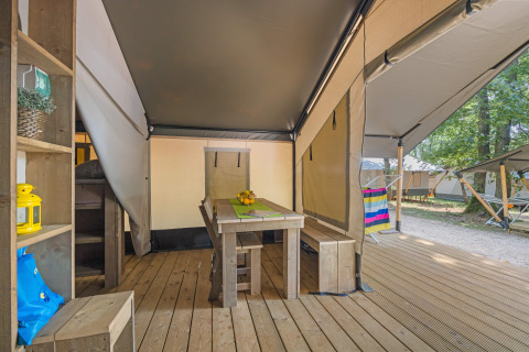 Intérieur d'une tente safari avec table en bois, bancs, et vue sur la terrasse et la nature extérieure.