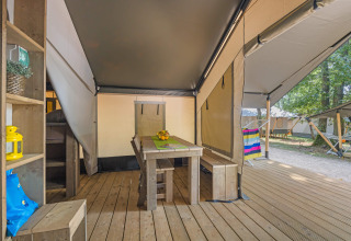 Intérieur d'une tente safari avec table en bois, bancs, et vue sur la terrasse et la nature extérieure.