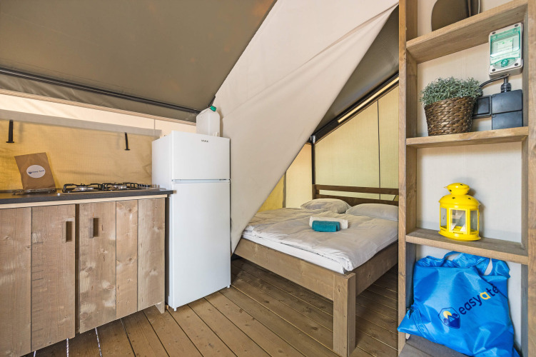 Intérieur d'une tente safari avec mobilier en bois, kitchenette, réfrigérateur et lit double sur plancher.