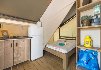 Interior of a safari tent featuring wooden furniture, kitchenette, fridge, and a double bed on wood floor.