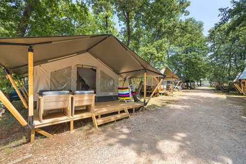 Tenda safari con veranda in legno e sedie in un campeggio alberato, altre tende lungo il sentiero.