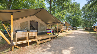 Tenda safari con veranda in legno e sedie in un campeggio alberato, altre tende lungo il sentiero.