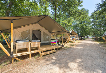 Tienda safari con terraza de madera y sillas en un camping arbolado, junto a un sendero de grava.