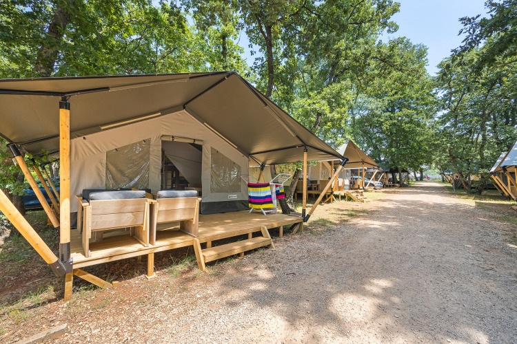 Tente safari avec terrasse en bois et fauteuils dans un camping boisé, d'autres tentes au fond.