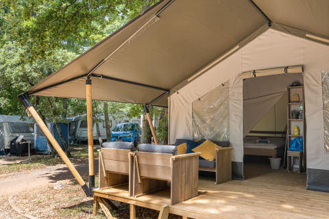 Tienda safari con terraza de madera, asientos exteriores y cama visible en un entorno de camping arbolado.
