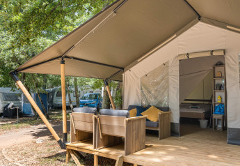 Tienda safari con terraza de madera, asientos exteriores y cama visible en un entorno de camping arbolado.