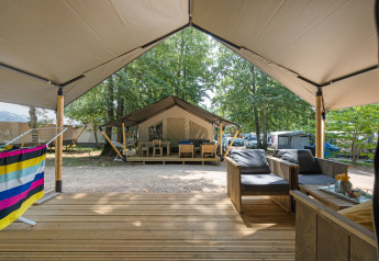 Udsigt fra en safaritelts terrasse med udendørs siddeområde og andre telte i en skovfyldt campingplads.