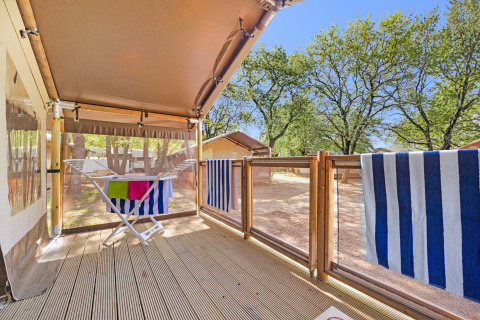 Veranda in legno con stendino e asciugamani a righe al Safari Tent Lodge tra alberi e cielo sereno.