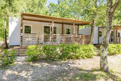 Vista del Mobile home Fuskulin, casetta moderna con veranda in legno, immersa nel verde tra alberi e cespugli.