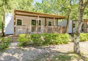 Vista del Mobile home Fuskulin, casetta moderna con veranda in legno, immersa nel verde tra alberi e cespugli.