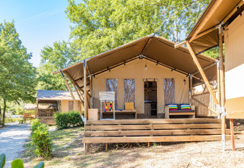 Tente safari avec terrasse, climatisation et sanitaires privés, entourée d’arbres et de verdure.