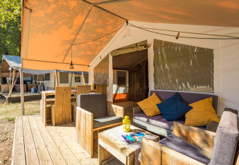 Safari Tent Luxury con veranda in legno, comode poltrone e cuscini colorati in un ambiente naturale.