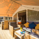 Safari Tent Luxury con veranda in legno, comode poltrone e cuscini colorati in un ambiente naturale.