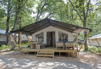 Vista esterna di una tenda safari con servizi igienici, terrazza in legno e area relax in una foresta.