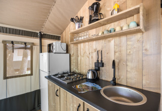Zona de cocina en tienda safari con paneles de madera, cocina de gas, fregadero y nevera en Camping Aminess Maravea.