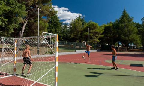 Jugendliche spielen Fußball auf einem Außenplatz im Koversada Camping Resort, umgeben von Bäumen.
