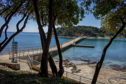 Muelle y costa rocosa vistos entre árboles en Koversada Camping Resort, Istria, Croacia, día soleado.