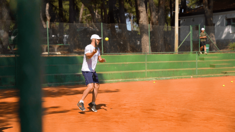 Hombre jugando tenis en una cancha de arcilla en Camping Porto Sole, Istria, Croacia, día soleado y árboles.