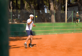 Mand spiller tennis på rød grusbane i Camping Porto Sole, Istrien, Kroatien. Solskinsdag omgivet af træer.