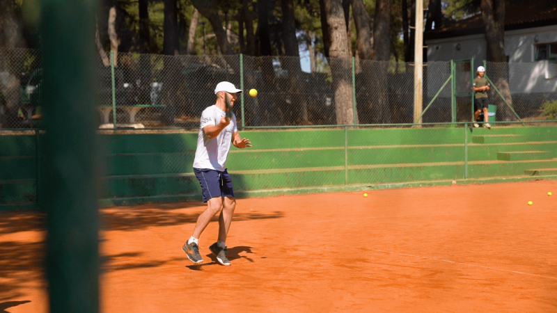 Man playing tennis on clay court at Camping Porto Sole, Istria, Croatia, surrounded by trees on a sunny day.