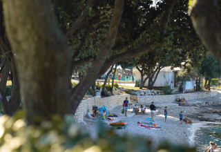 Persone si rilassano sulla spiaggia di ciottoli al Camping Porto Sole, Istria, Croazia, tra alberi frondosi.