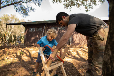 Un hombre ayuda a un niño a serrar madera al aire libre en Feather Down Figari Corsica, parque vacacional en Francia.