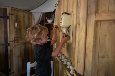 Een kind gebruikt een wandgemonteerde koffiemolen in een houten hut op Feather Down Figari Corsica vakantiedomein.