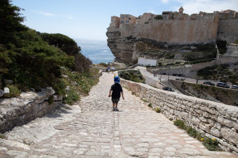 A child walks down a cobblestone path near Figari, Corsica, France, overlooking the sea and dramatic cliffs.
