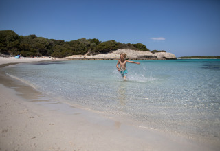 A child runs through shallow, clear water at a sandy beach near Figari in sunny Corsica, France.