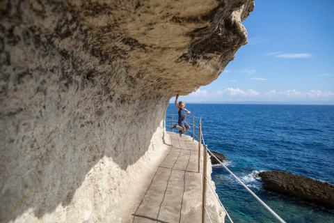 Une personne saute et touche la falaise sur un chemin étroit surplombant la mer bleue en Corse, France.