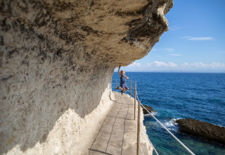 Una persona salta e tocca la scogliera lungo un sentiero stretto affacciato sul mare blu in Corsica, Francia.