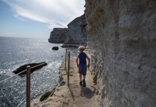 Dreng går på en smal kyststi udhugget i klippen nær Figari, Corsica, med havet til venstre og solskin.