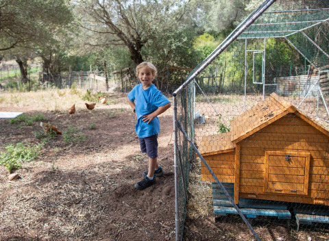 Niño con camiseta azul sonríe junto a gallinero en Feather Down Figari Corsica, parque vacacional en Francia.