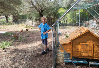 Niño con camiseta azul sonríe junto a gallinero en Feather Down Figari Corsica, parque vacacional en Francia.