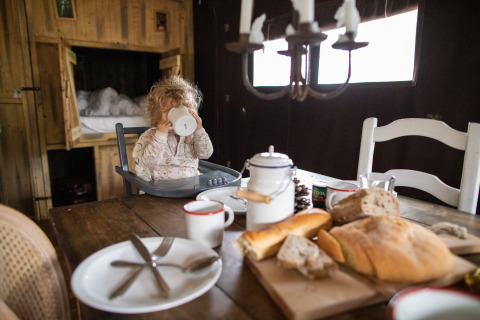 Small child in high chair drinks from a mug at a rustic breakfast table with bread in Corsica, France.
