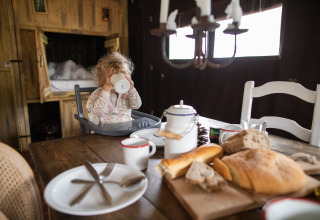 Small child in high chair drinks from a mug at a rustic breakfast table with bread in Corsica, France.