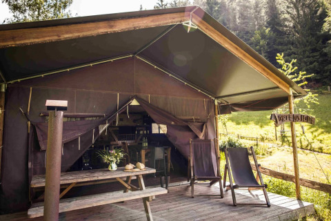 Tienda tipo safari con terraza de madera, dos tumbonas y mesa de picnic, rodeada de naturaleza y bosque.