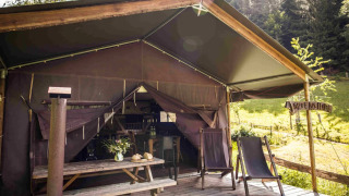Safari tent with a wooden deck, two lounge chairs, and picnic table, surrounded by greenery and forest.