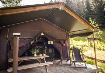Tenda safari con veranda in legno, due sedie a sdraio e tavolo da picnic, circondata dal verde.