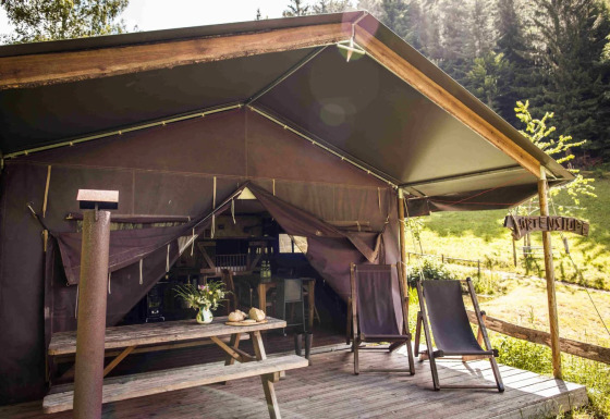 Tienda tipo safari con terraza de madera, dos tumbonas y mesa de picnic, rodeada de naturaleza y bosque.