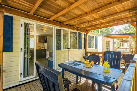 Overdekt terras van Mobile home Capri met plastieken tafel, stoelen en zicht op de keuken binnen.