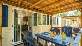 Mobilhome Capri udendørs terrasse med spisebord, stole og åben adgang til køkken under trætelt.