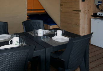 Zona pranzo in tenda safari con tavolo apparecchiato, sedie nere e letto, Minicamping de Vrolijke Flierefluiter.