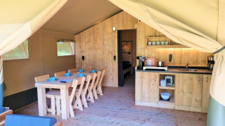 Interior of a luxury safari tent with wooden furniture, dining table, kitchen, and a view of the bedroom.