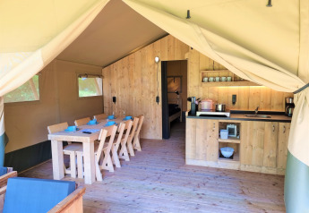 Interieur van een luxueuze safaritent met houten meubels, eettafel, keuken en zicht op de slaapkamer.