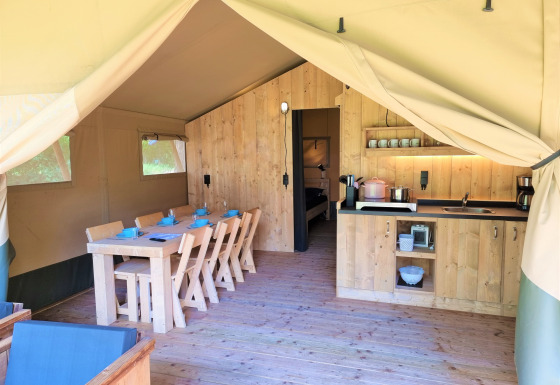 Interno di una tenda safari di lusso con mobili in legno, tavolo da pranzo, cucina e vista sulla camera.