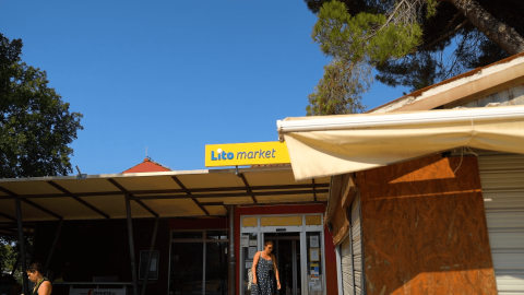 Una mujer sale de Lito Market en Camping Polari, Istria, Croacia, con cielo azul y árboles de fondo.